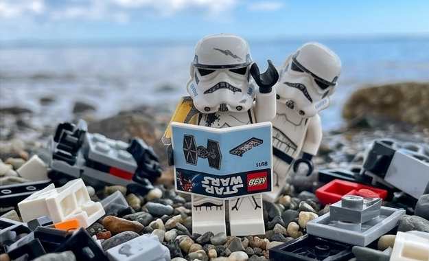 A LEGO stormtrooper holds a tiny printed manual upside down, scratching his helmet, while a second trooper leans in, just as lost.
